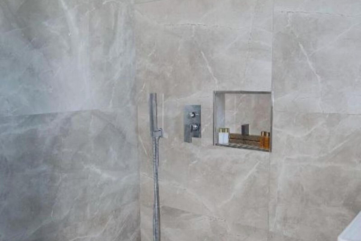 A bathroom with a marble wall, a shower with a handheld showerhead, and a small shelf with toiletries.