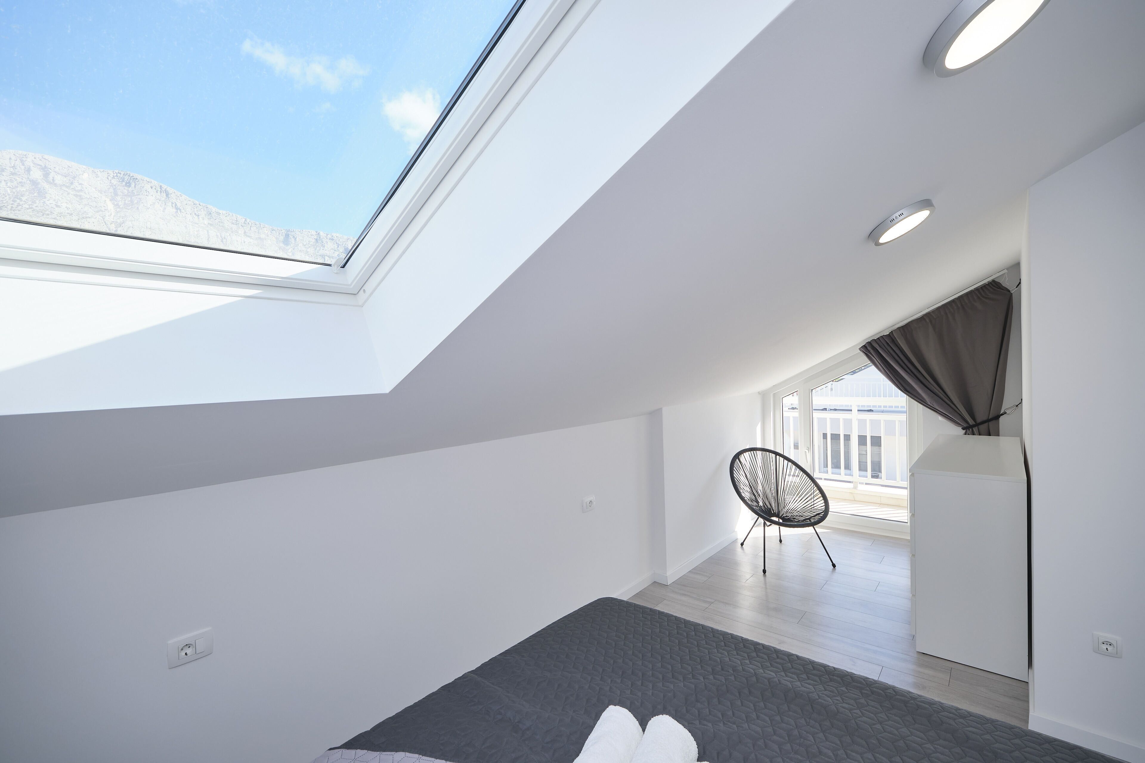 A modern bedroom with a skylight, a bed, a chair, and a view of the outdoors.