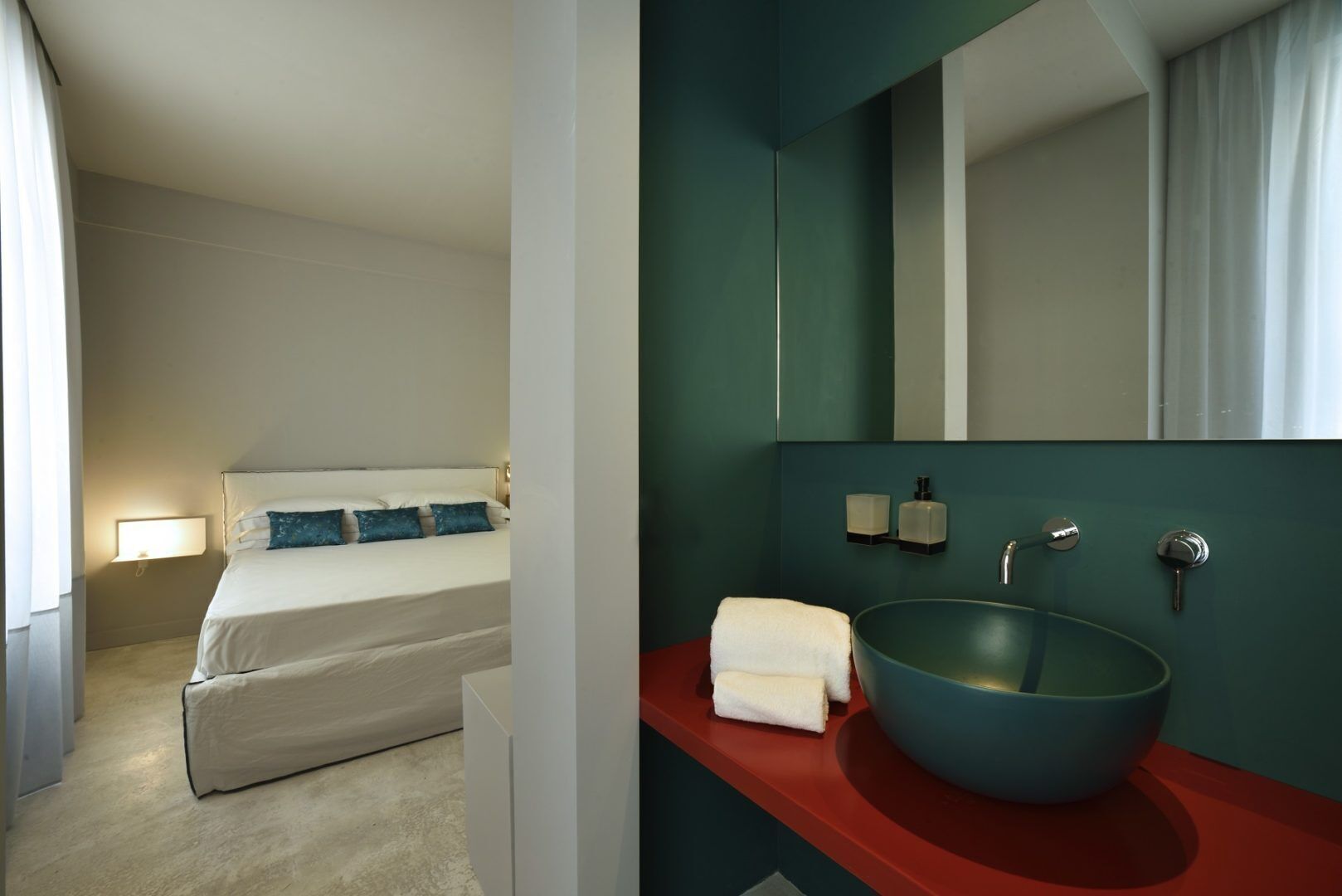 Habitación de hotel con una cama, una mesita de noche con lámpara, una encimera roja con lavabo y espejo, y un tazón azul.