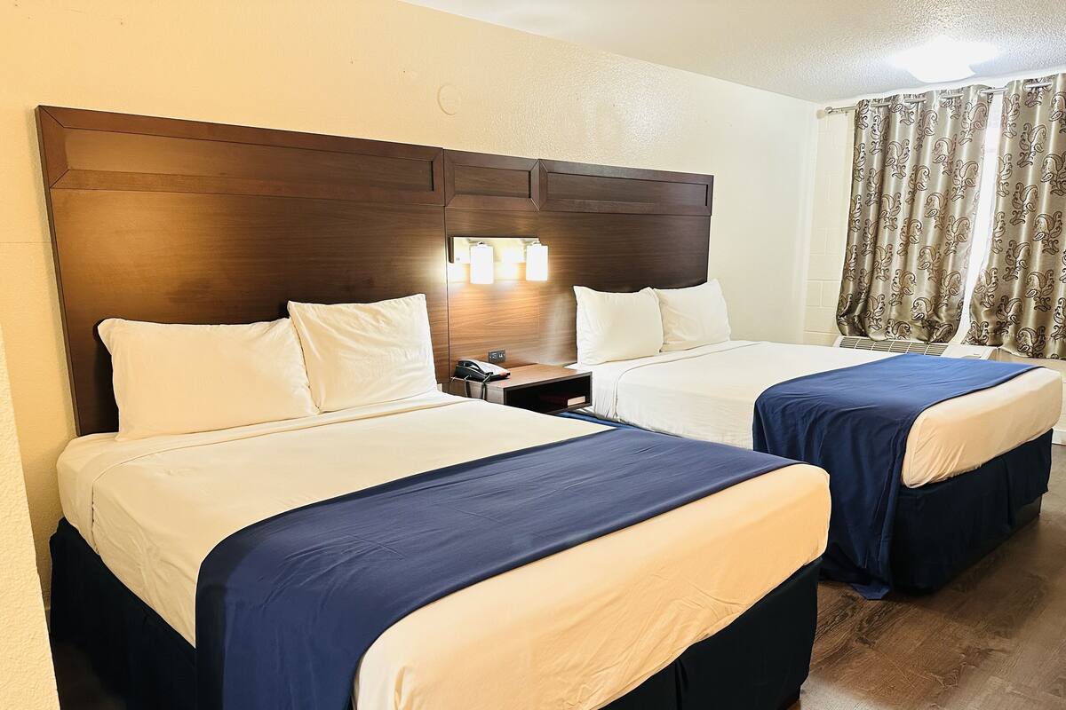 Habitación de hotel con dos camas, cada una con ropa de cama blanca y azul, cabeceros de madera, mesitas de noche con lámparas y cortinas con estampado paisley.