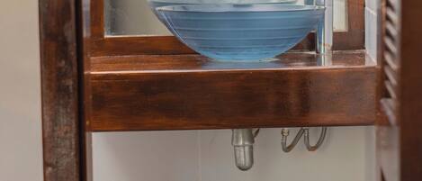 A bathroom with a wooden vanity, a blue vessel sink, a mirror, and a wooden waste basket.