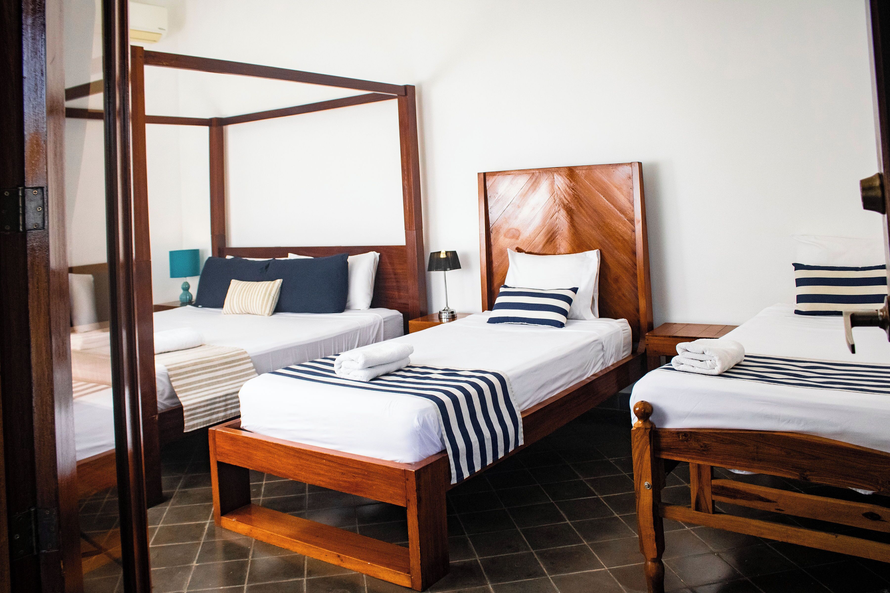 Habitación de hotel con dos camas individuales, cabecera de madera, mesita de noche con lámpara y vistas a través de una puerta abierta.