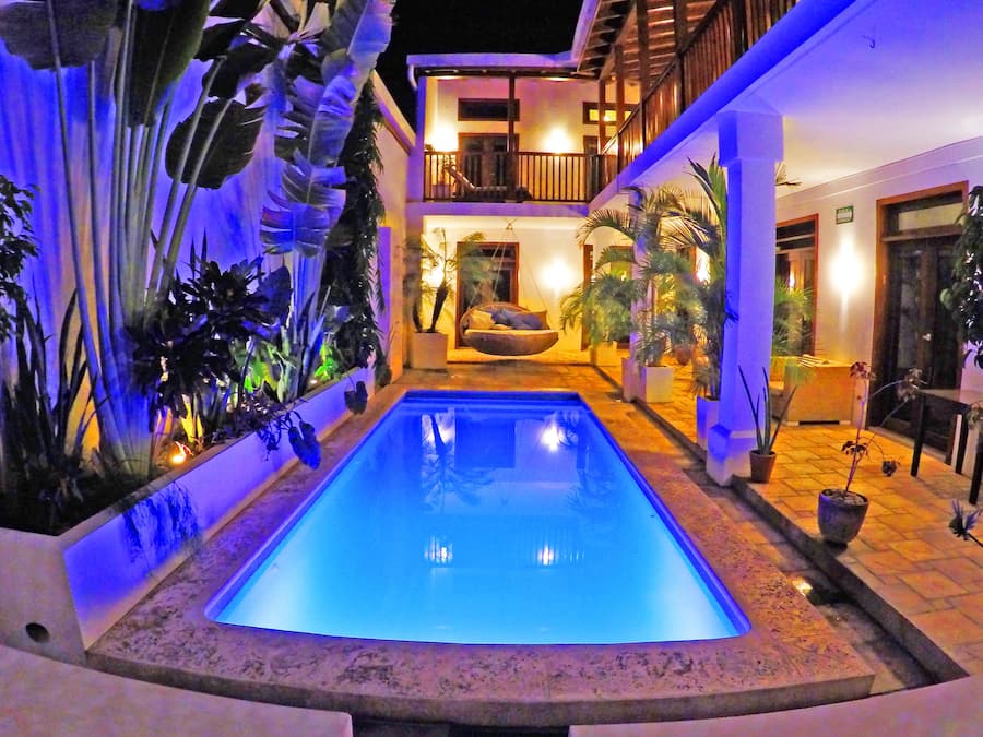 Una piscina con borde de piedra, rodeada de plantas en macetas y un edificio de dos pisos con balcones.
