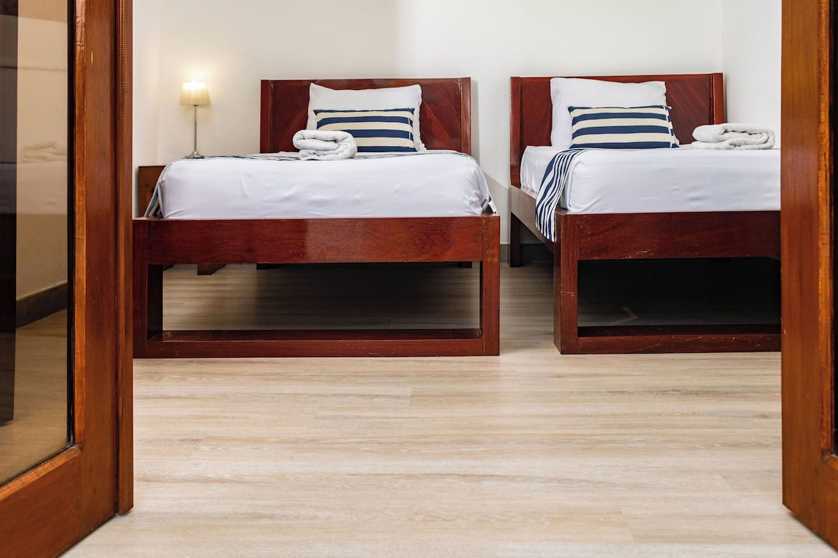 Duas camas de madeira com lençóis brancos e almofadas listradas, um abajur e piso de madeira.