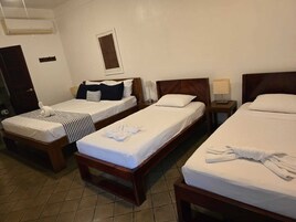 Habitación de hotel con tres camas individuales, cada una con cabecera de madera y ropa de cama blanca. Hay un cuadro enmarcado en la pared sobre las camas, una mesita de noche con lámpara junto a la cama central y una puerta a la izquierda.