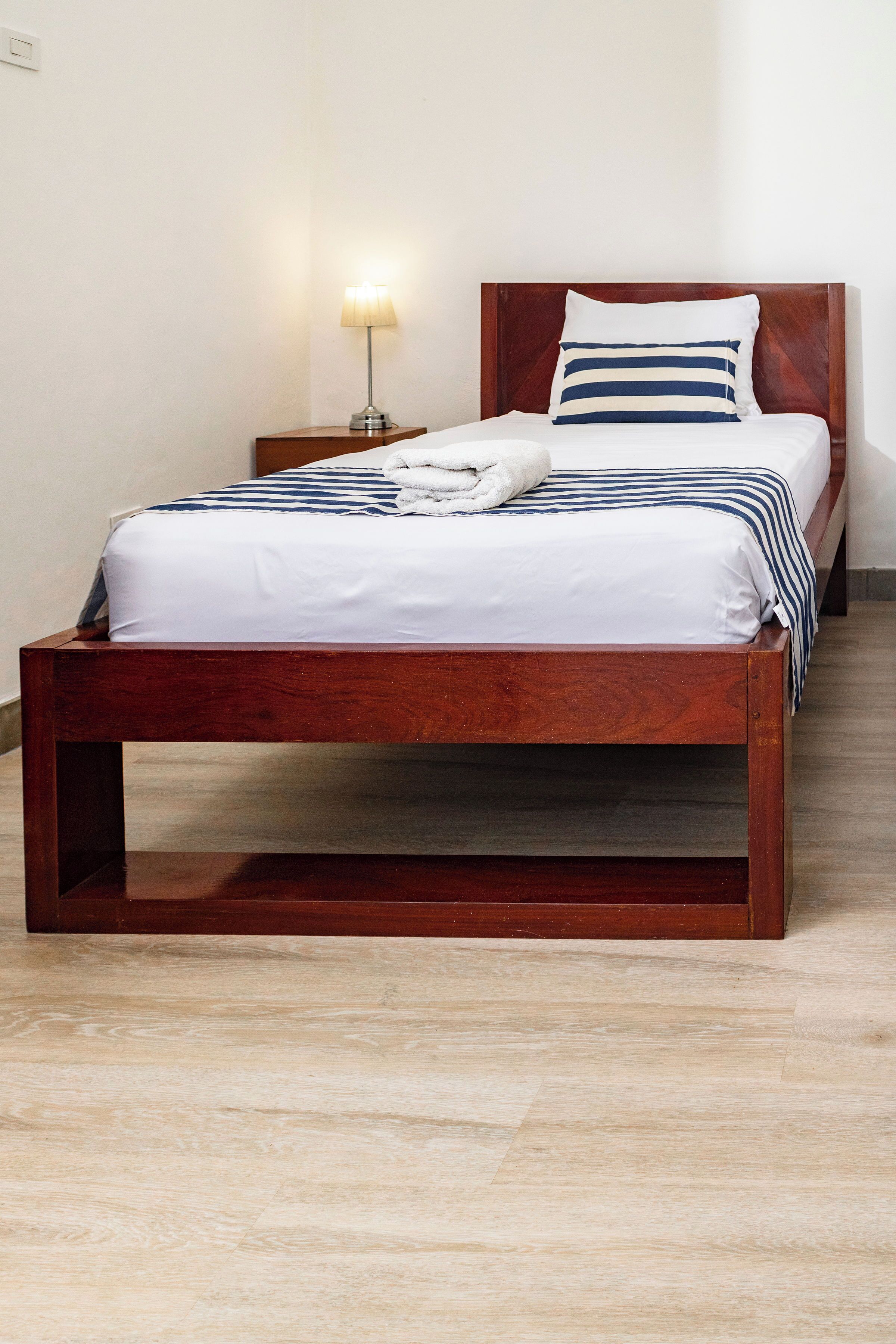 A wooden bed with a white and blue striped bedspread, a wooden nightstand with a lamp, and a wooden floor.