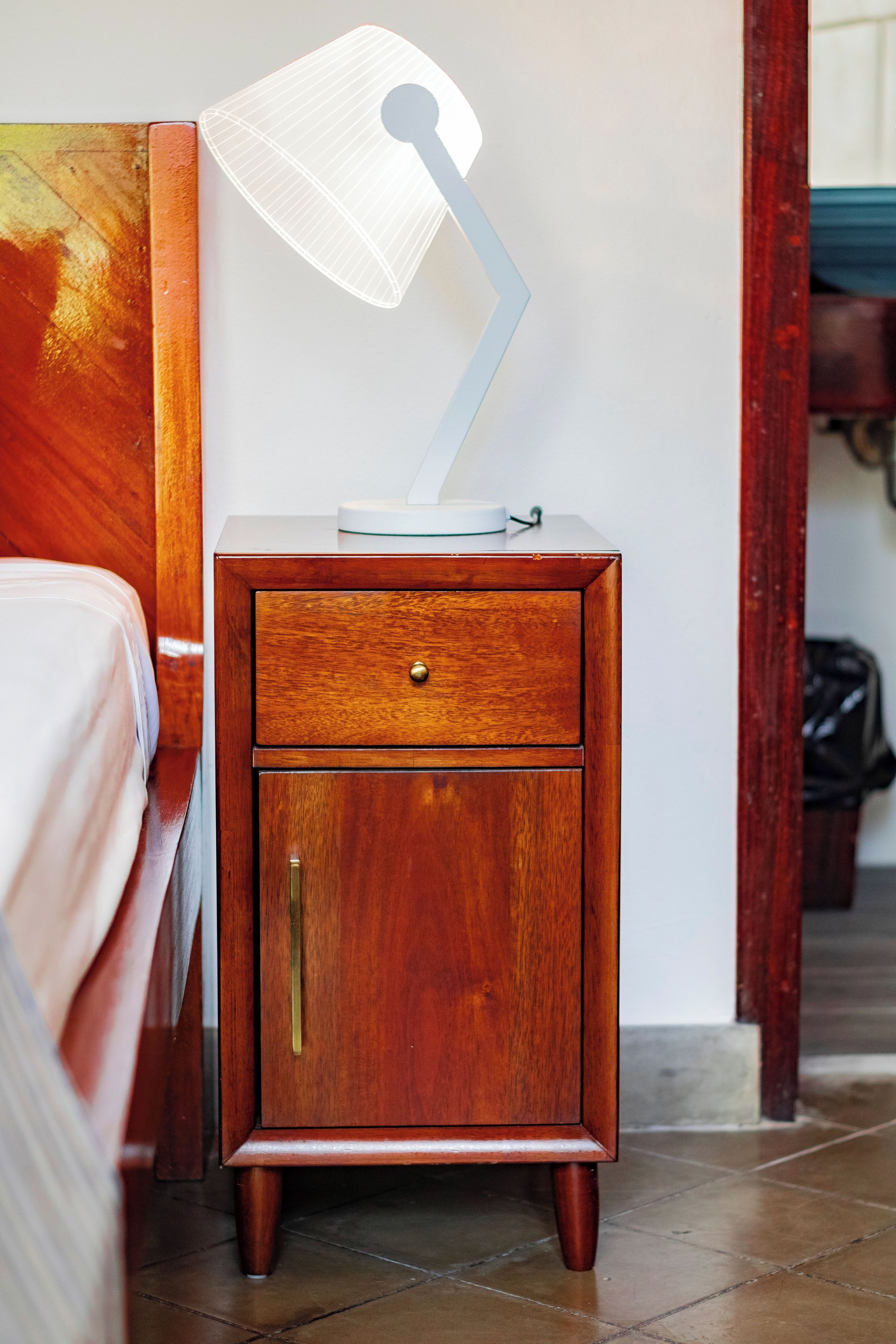 Een houten nachtkastje met een lade, een lamp en een klein tafeltje met een wit voorwerp erboven.