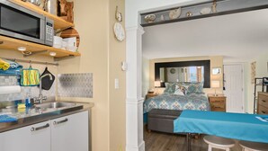 Een kleine keuken met een magnetron, spoelbak en opslagplanken. Achter een open deur is een slaapkamer te zien met een bed, nachtkastjes en een grote spiegel.