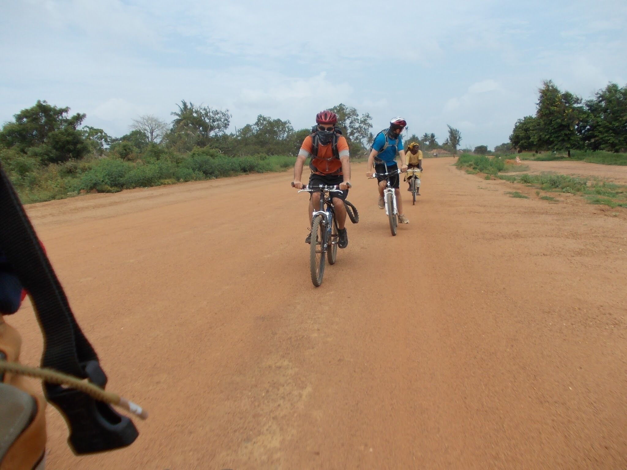 Three cyclists on a dirt road with sparse vegetation and trees in the background.