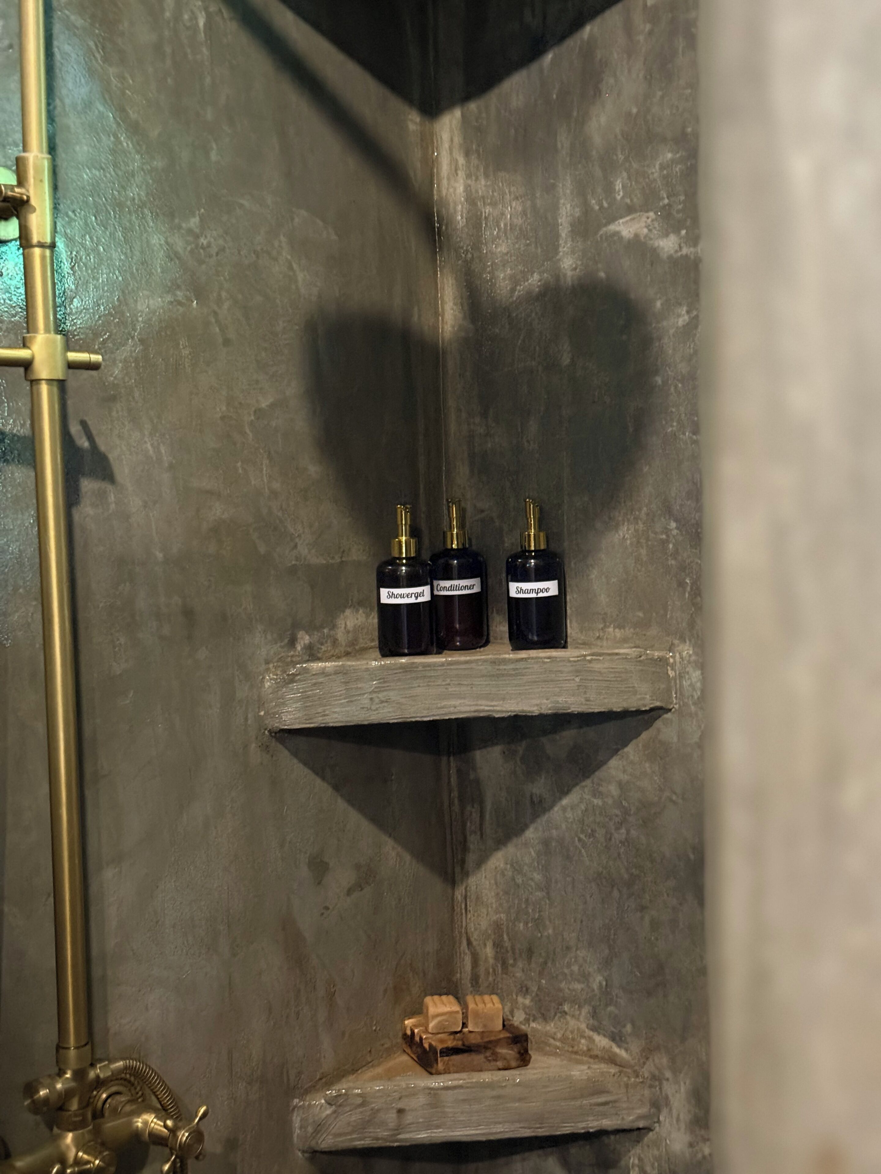 Un baño con zona de ducha, un estante con tres botellas etiquetadas como 'Showergel', 'Acondicionador' y 'Champú', y un bloque de madera.