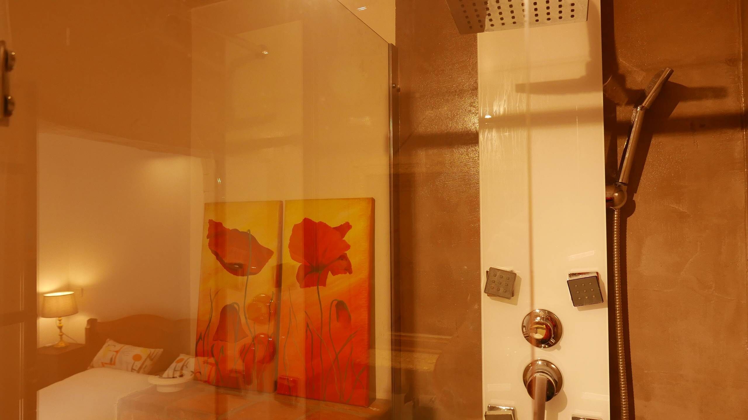 A hotel bathroom with a glass shower enclosure, a bed with a nightstand, and a wall-mounted light.