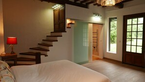 A bedroom with a wooden staircase, a bed, a nightstand with a lamp, and a large window with a view of greenery. - Villa Lion-Mallet (MIANNAY)