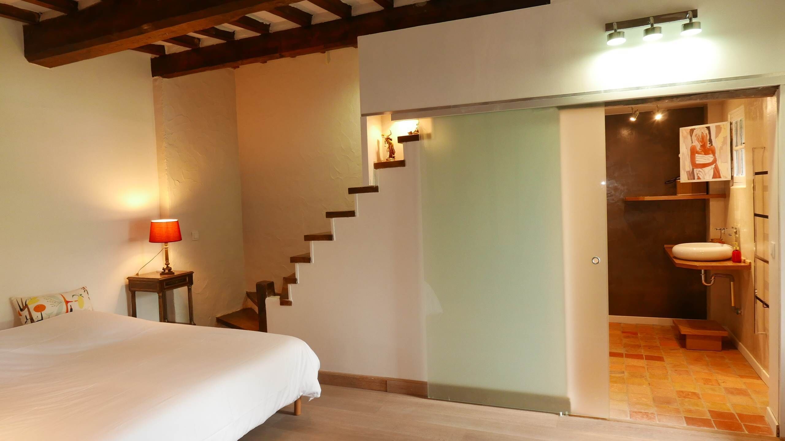A bedroom with a bed, a nightstand with a lamp, a staircase, and a bathroom with a sink and towel rack.