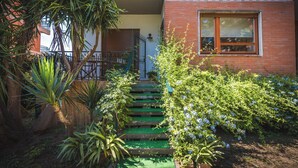 A two-story building with a green staircase leading to the entrance, surrounded by lush greenery and a small balcony.