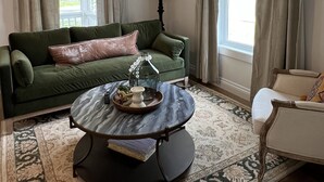 En stue med en grøn sofa, et rundt sofabord, en lampe og udsigt ud af et vindue med gardiner.