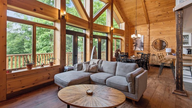 Eine geräumige Holzhütte mit großzügigem Wohnbereich, ausgestattet mit einer Eckcouch, einem runden Couchtisch und großen Fenstern mit Blick auf den umliegenden Wald.
