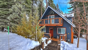 A two-story cabin with a wooden deck and a red door, surrounded by snow and trees. - Cozy Cabin on Brule - Hot Tub, Jungle Gym & Games (South Lake Tahoe)