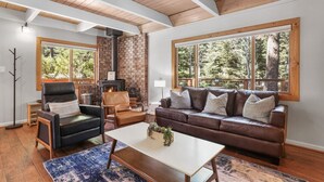 A cozy living room with a fireplace, leather furniture, and large windows offering a view of the forest. - Cozy Cabin on Brule - Hot Tub, Jungle Gym & Games (South Lake Tahoe)