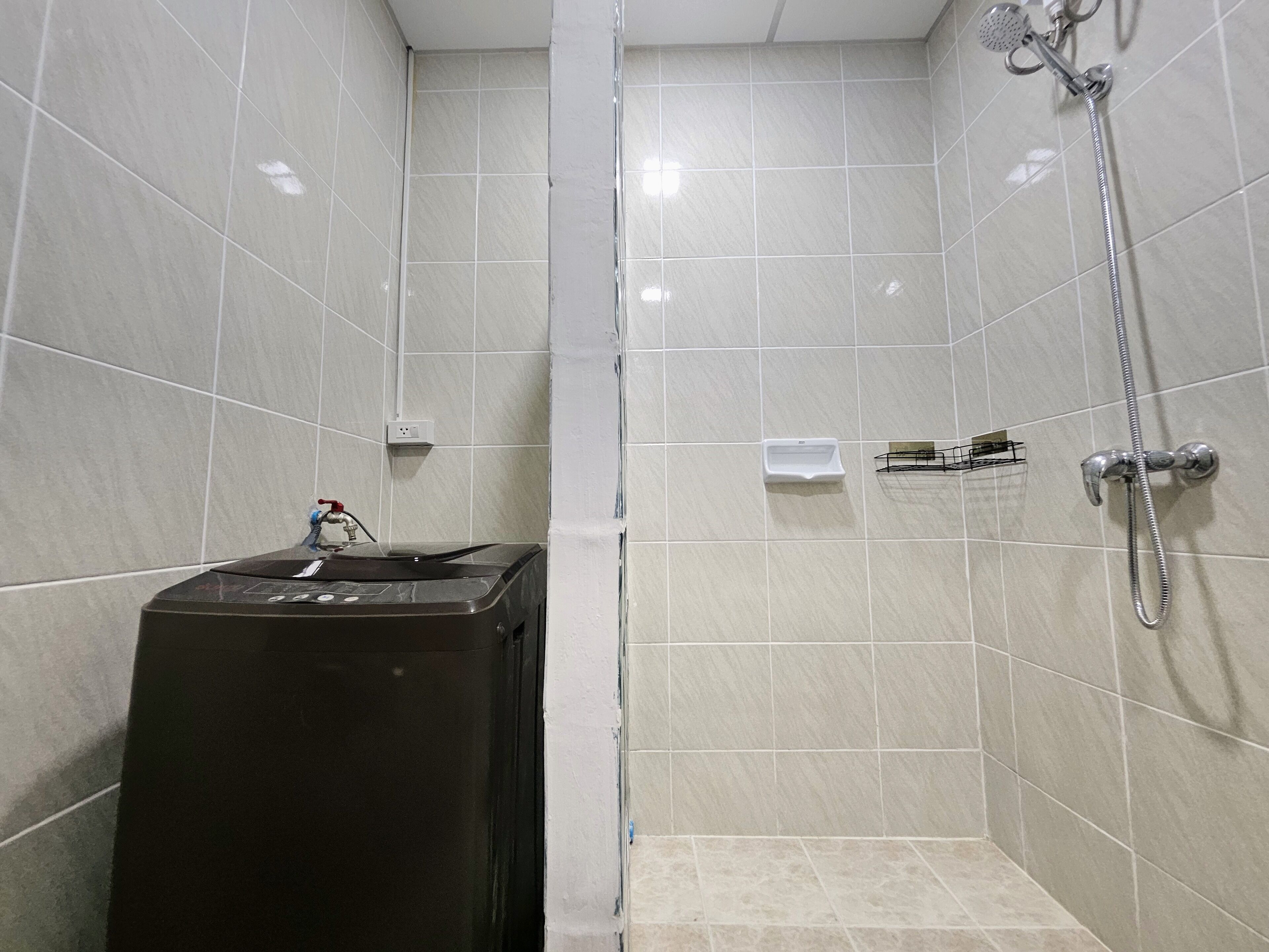 Un espacio de ducha pequeño con azulejos, un cabezal de ducha de mano, un dispensador de jabón y un objeto negro que parece un calentador portátil.