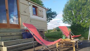Puinen terassi, jossa on kaksi punaista tuolia ja pieni pöytä, sininen, kudottu laukku ja puinen penkki.