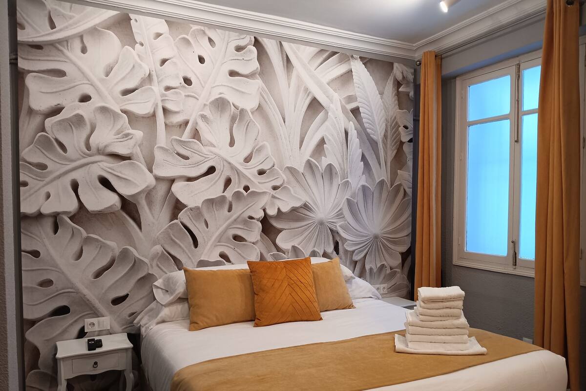 Uma cama bem arrumada com esquema de cores branco e marrom, uma mesa de cabeceira com abajur e uma janela com cortinas.
