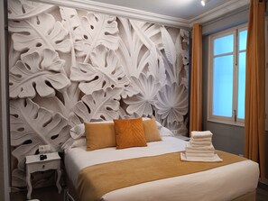 Ein ordentlich gemachtes Bett in Weißbraun, ein Nachttisch mit einer Lampe und ein Fenster mit Vorhängen.