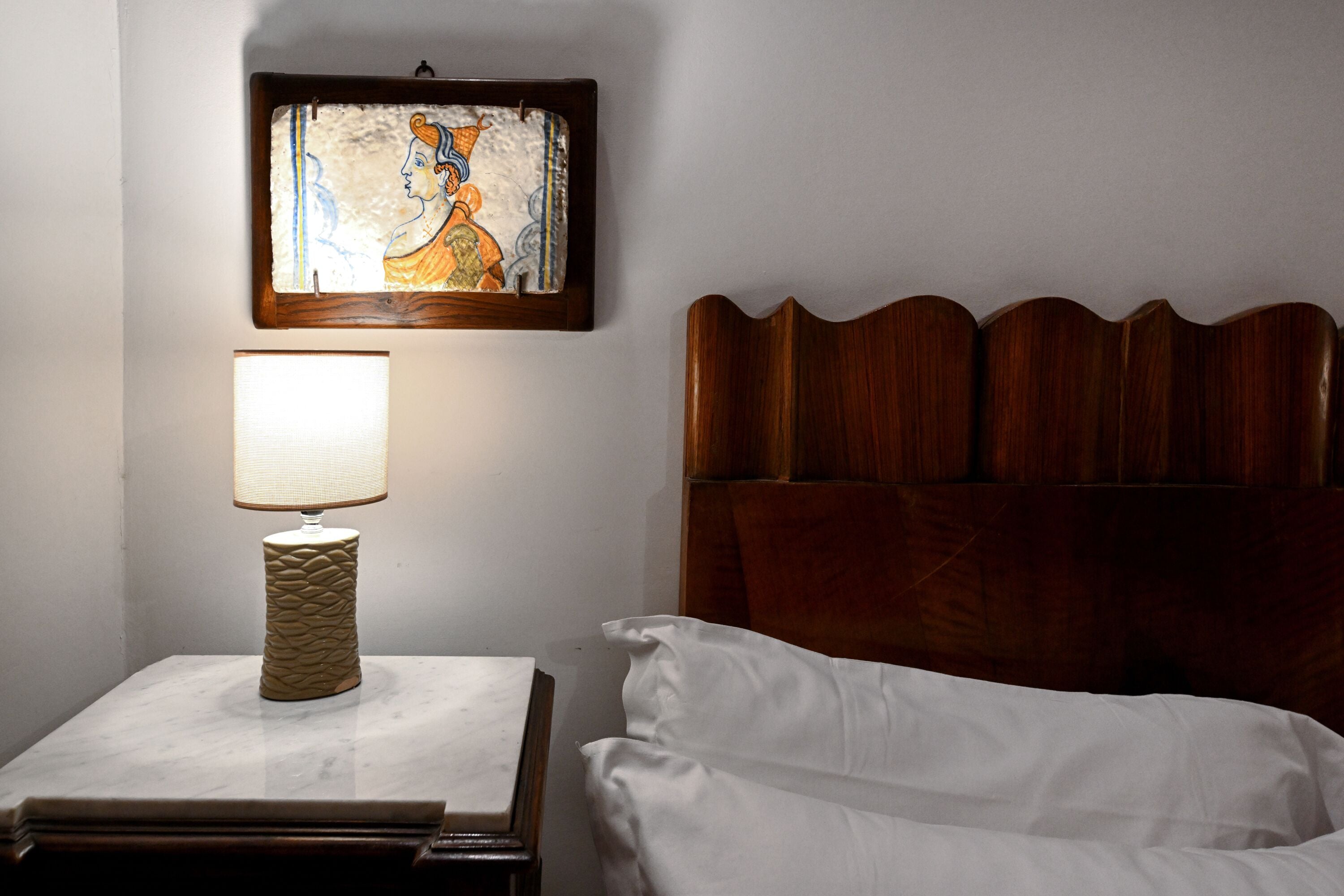 Un lit bien fait, avec du linge de lit blanc, une tête de lit en bois, une table de chevet avec une lampe et une œuvre d’art encadrée accrochée au mur.