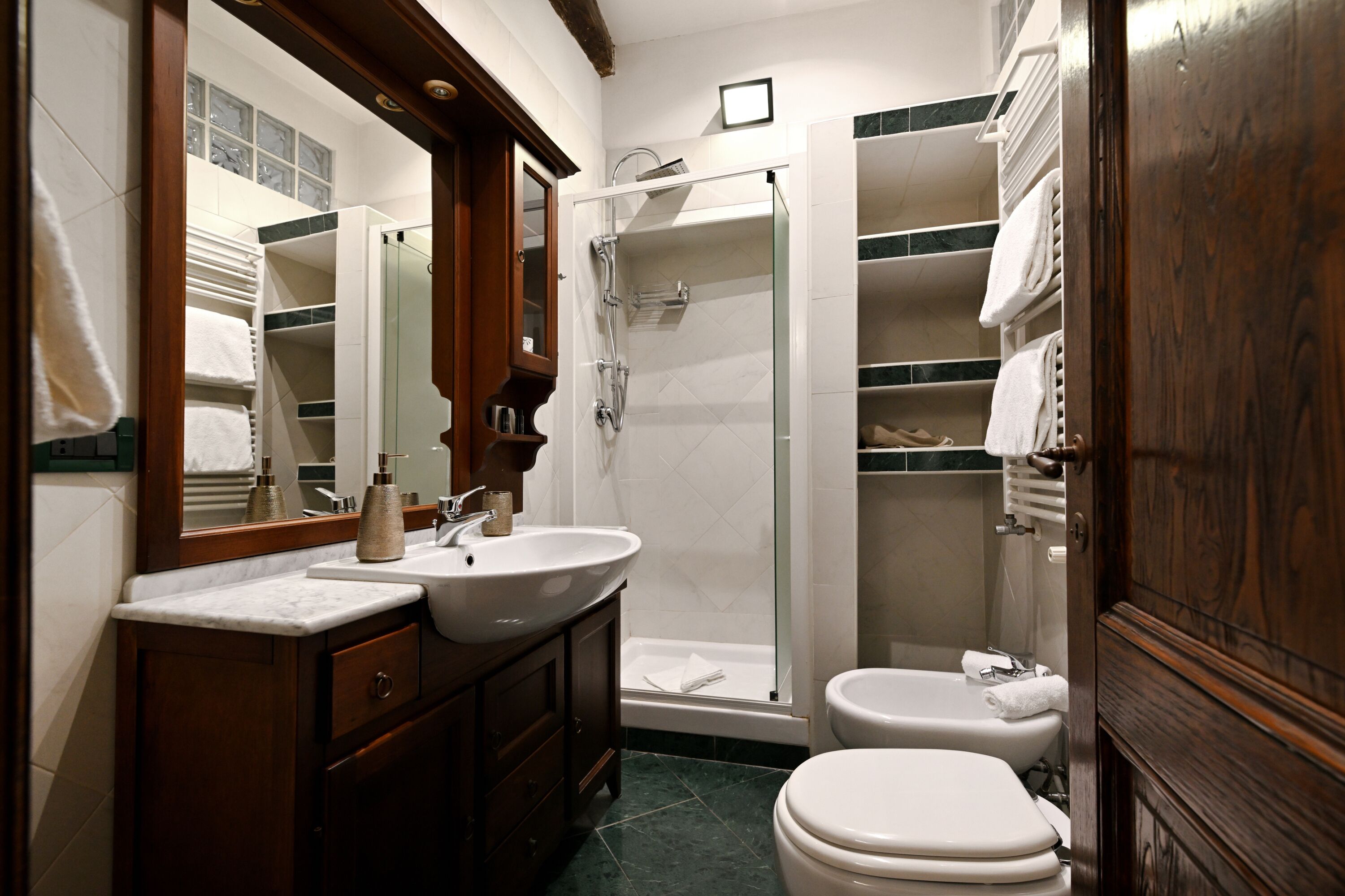 A bathroom with a double sink, a mirror, a shower, a toilet, and a built-in shelf with towels.