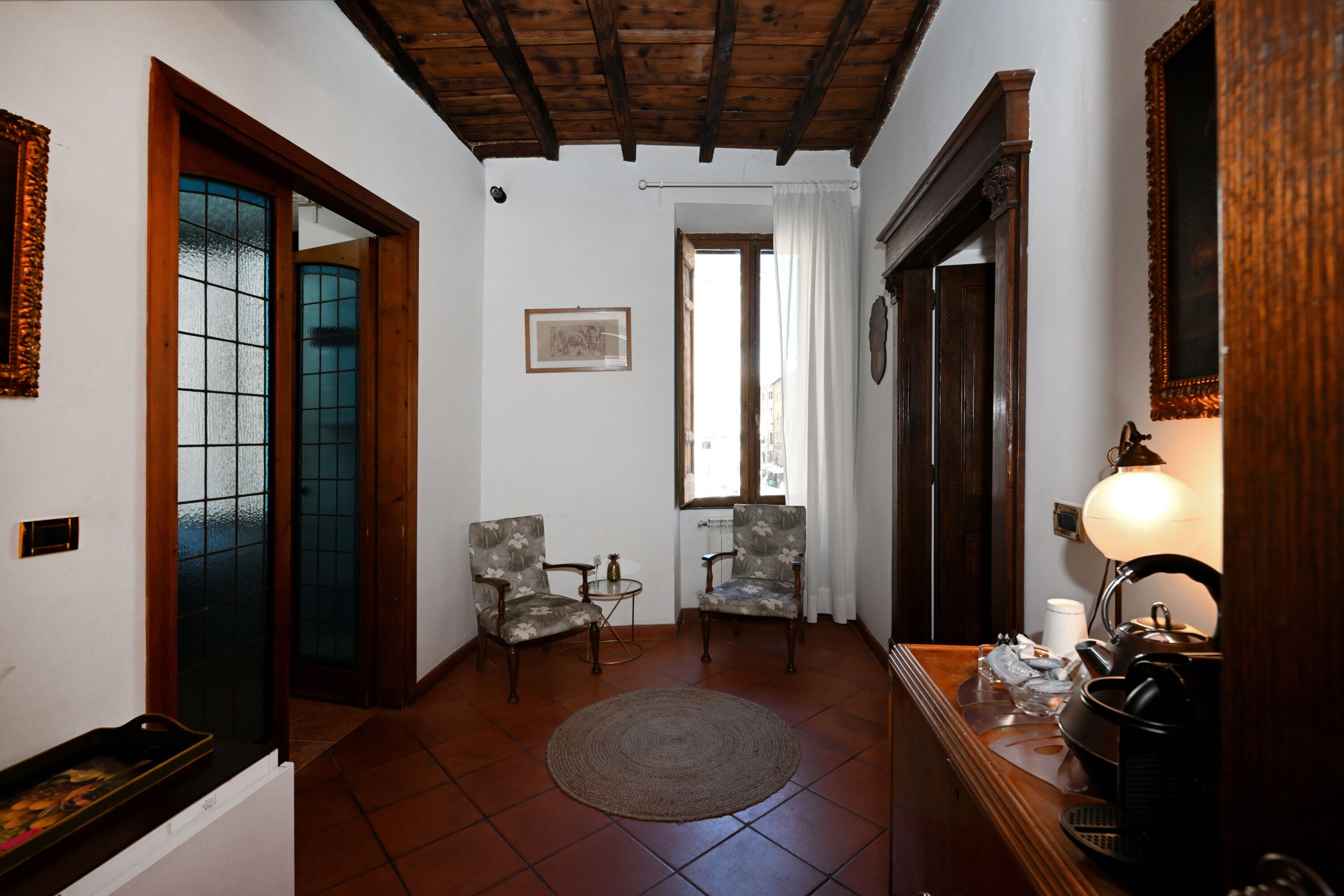 A room with a wooden ceiling, two chairs, a round rug, a table with a lamp, and a window with curtains.