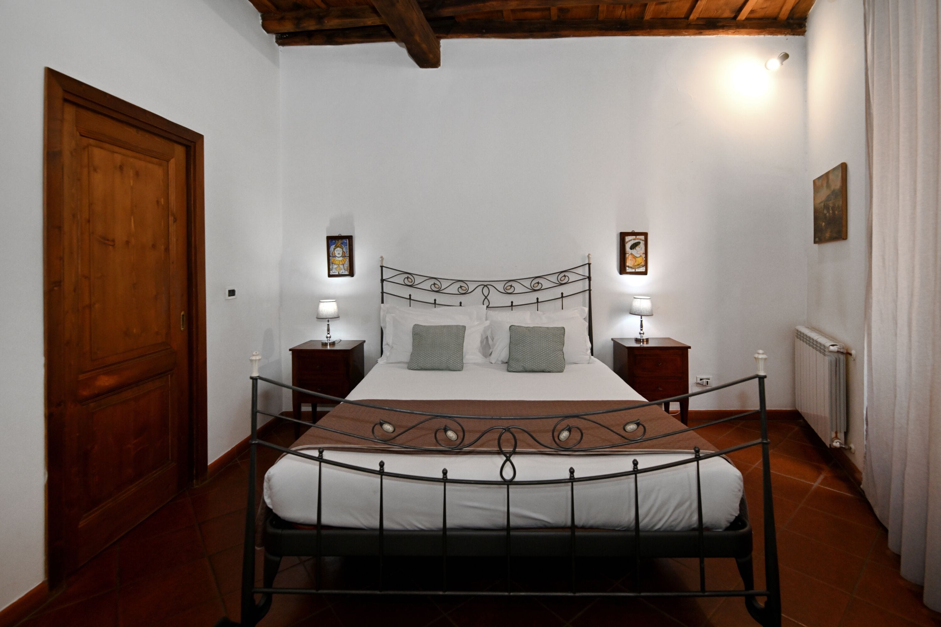 A bedroom with a metal bed frame, white bedding, two bedside tables with lamps, a wooden door, and a radiator.