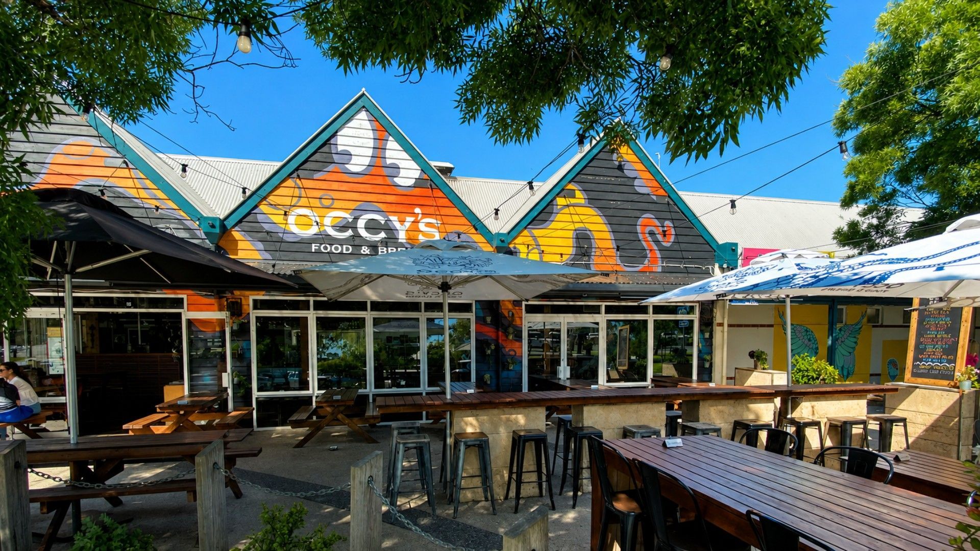 An outdoor seating area with tables and chairs, a covered patio, and a building with a colorful awning.