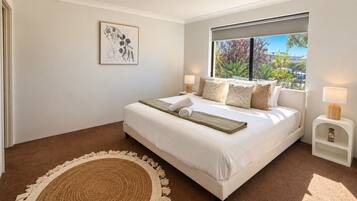 A bedroom with a large bed, a bedside table with a lamp, a window with a view of greenery, and a wall-mounted artwork.