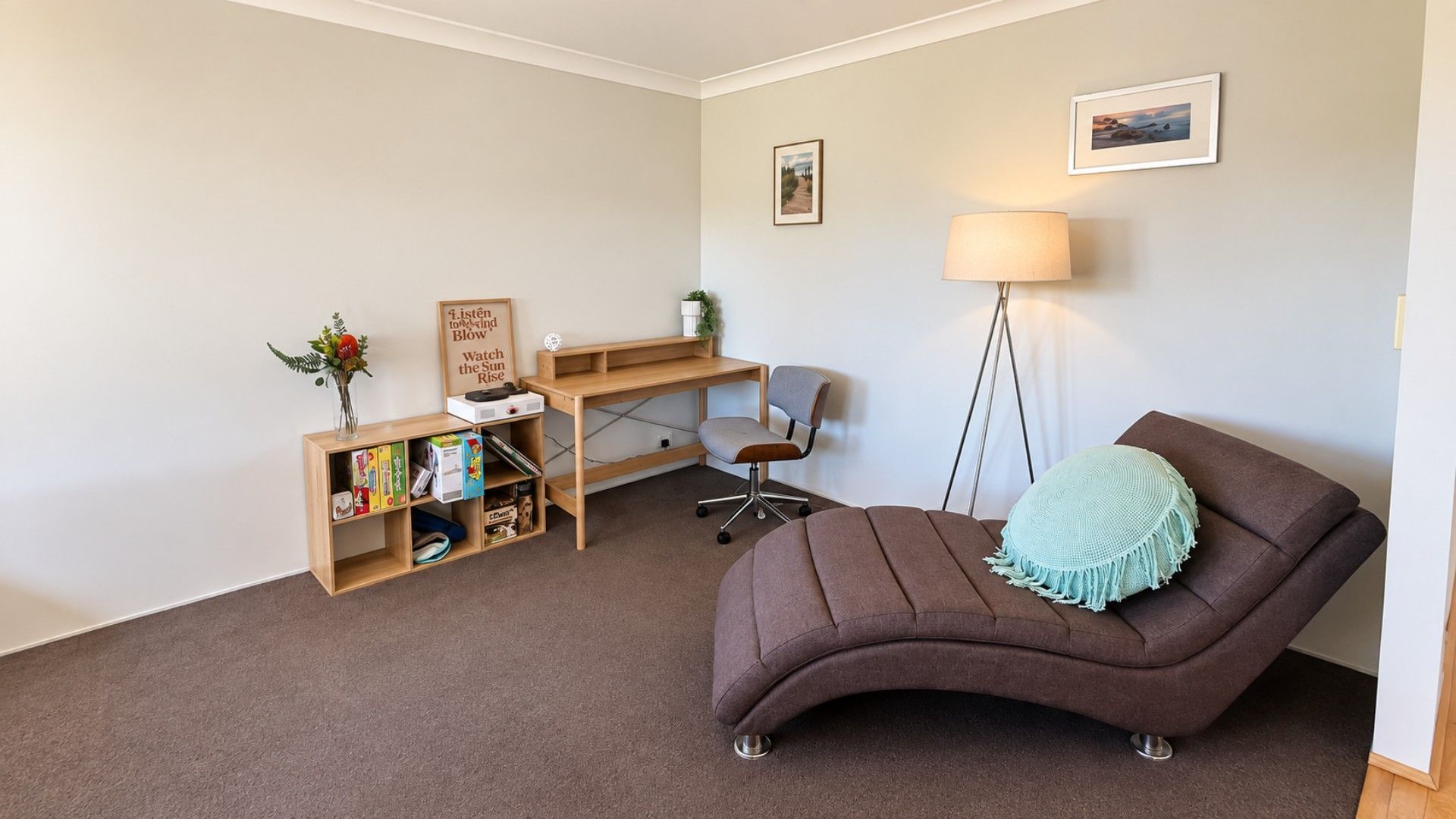 A room with a brown sofa, a desk with a chair, a bookshelf, a floor lamp, and two framed pictures on the wall.