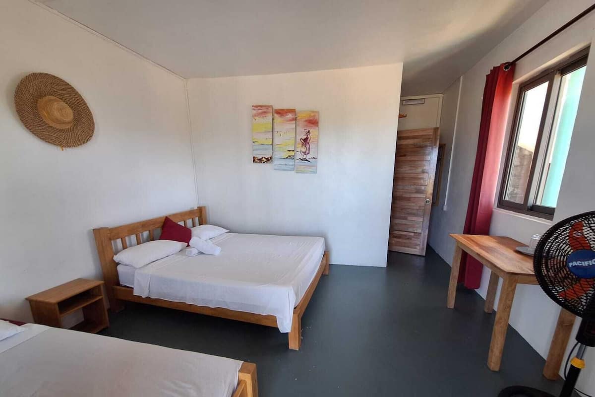 A room with two single beds, a wooden desk, a standing fan, and a straw hat on the wall.