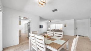 Un vaste espace dédié à la salle à manger, avec une table en bois et des chaises blanches, un ventilateur de plafond et un lustre moderne.