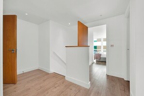 A hallway with wooden flooring and white walls, leading to a room with a bed and a window.