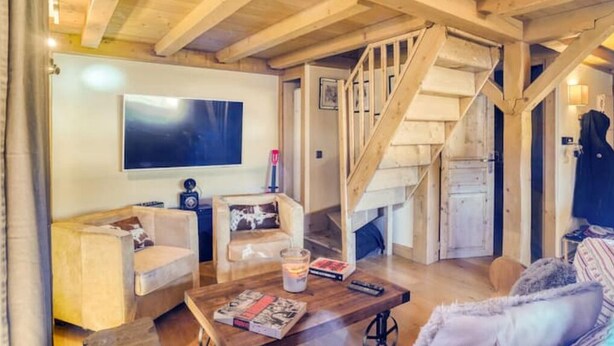 A cozy living room with a wooden staircase, a flat-screen TV, and a coffee table with books and a candle. - Le Wood Megève (Megève)