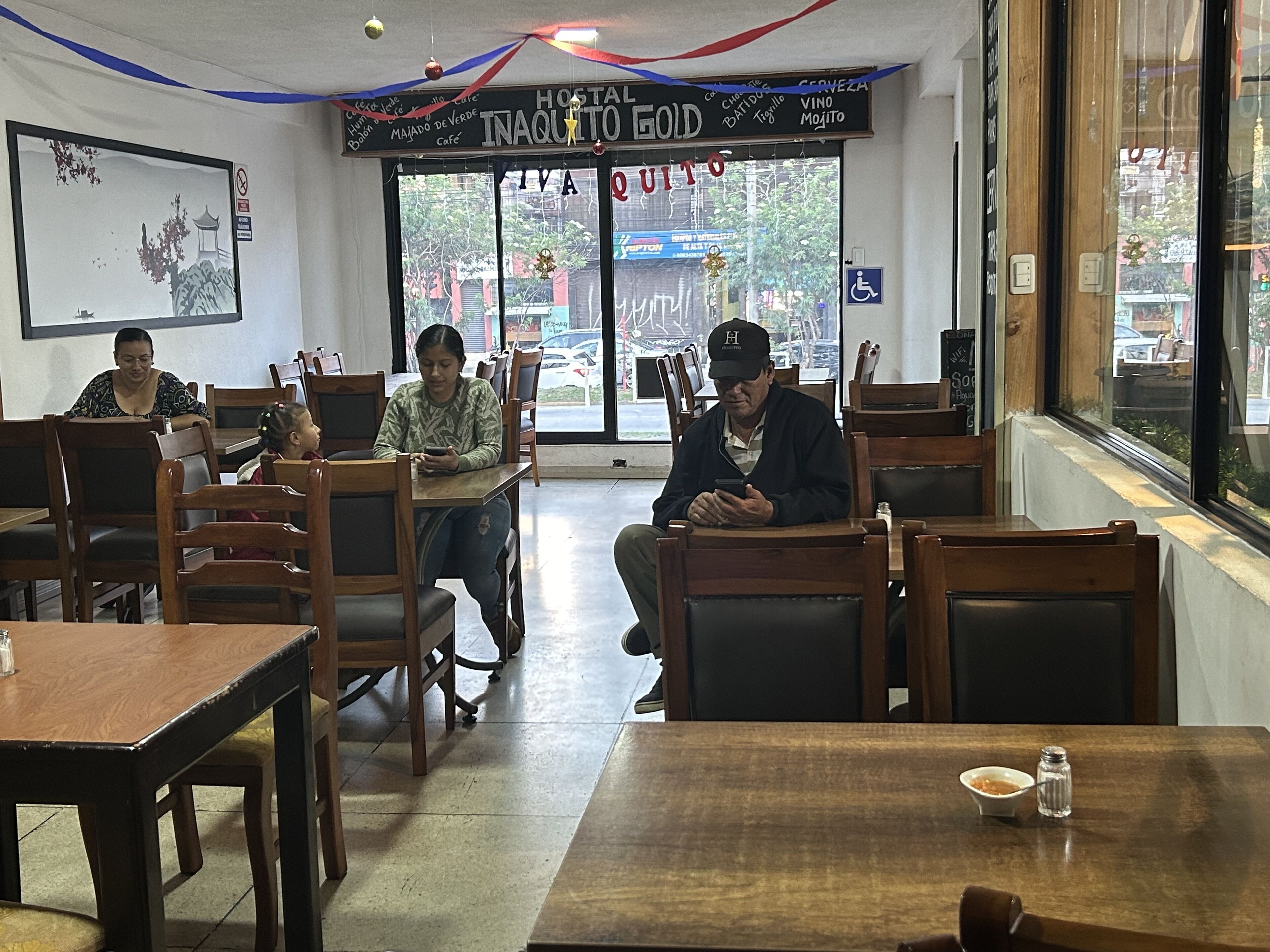 Interior de restaurante com mesas e cadeiras de madeira, uma janela grande com vista para a rua, e uma placa com a inscrição 