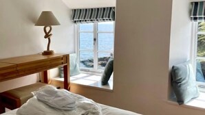 A bedroom with a bed, a wooden desk, a lamp, and a window with a view of the sea. - Cottages at the Lugger (Truro)