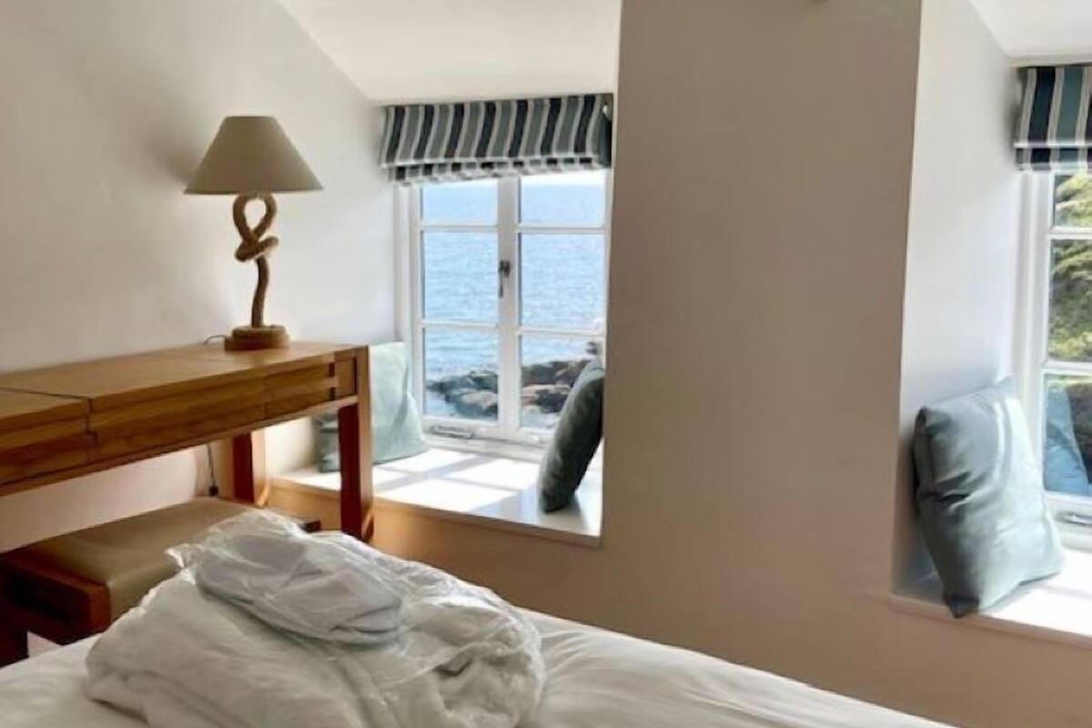 A bedroom with a bed, a wooden desk, a lamp, and a window with a view of the sea.