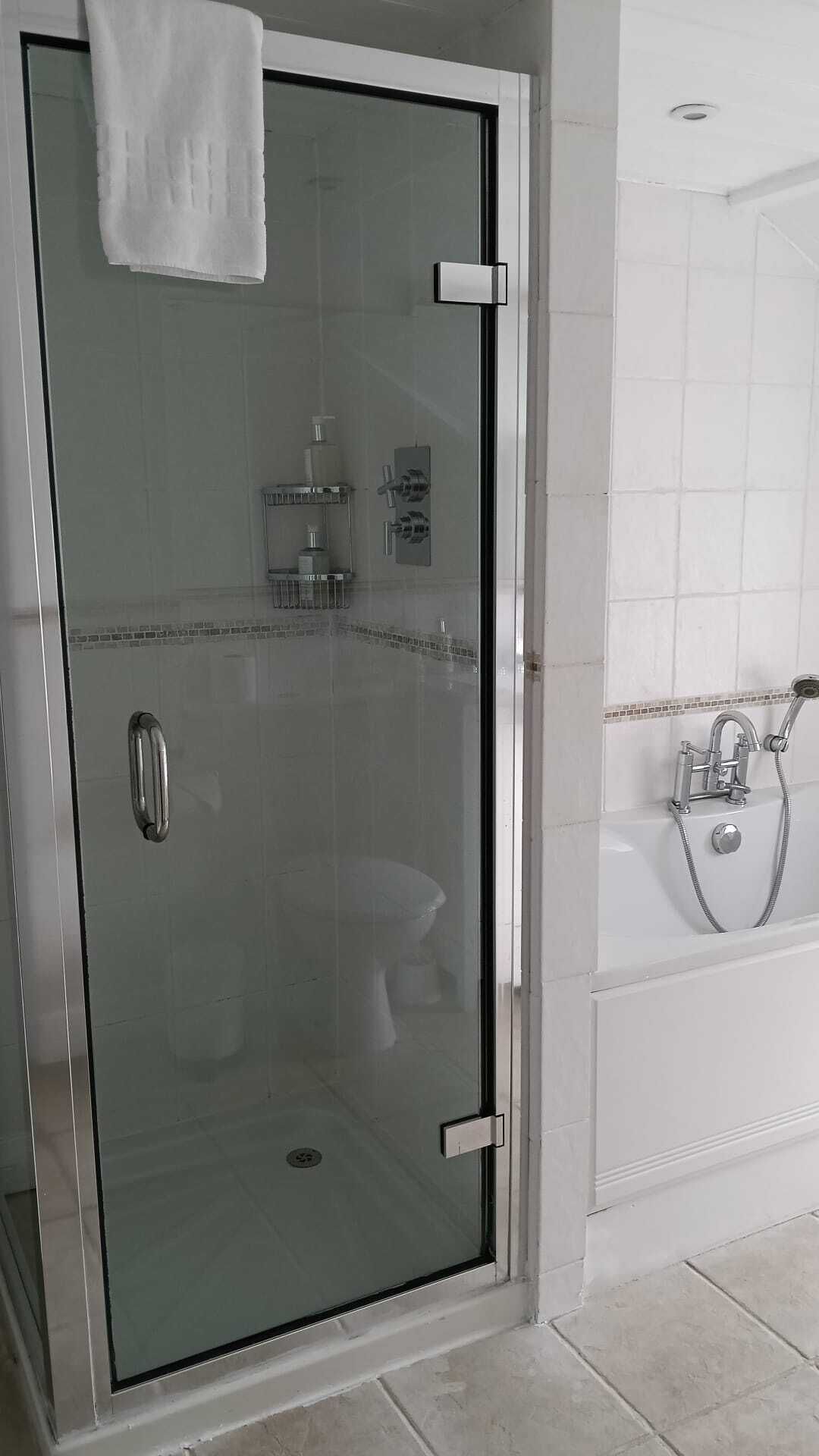 En brusekabine med glasdøre, et håndklæde hængende på døren, et toilet med glasvægge og et badekar med håndbruser.