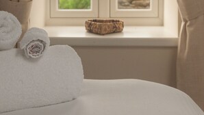 A bedroom with a bed, two rolled towels, a window with curtains, and a view of greenery and a building outside. - Cottages at the Lugger (Truro)