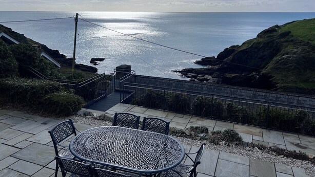 A patio with a round metal table and chairs, overlooking a coastal landscape with cliffs and the sea. - Cottages at the Lugger (Truro)