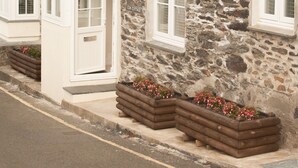 A stone cottage with white windows and doors, a sloped roof, and wooden flower beds in front. - Cottages at the Lugger (Truro)