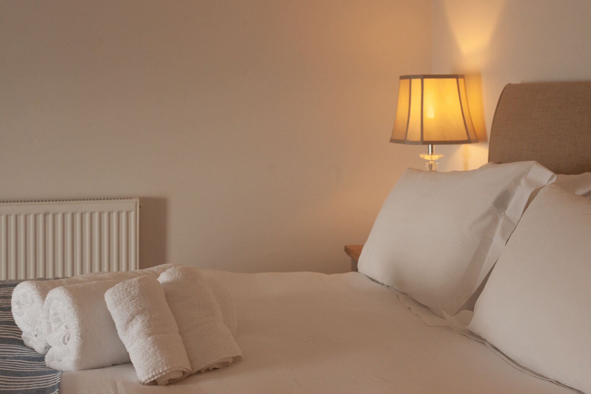 A neatly made bed with white linens and two rolled towels, a bedside lamp, and a radiator on the wall.