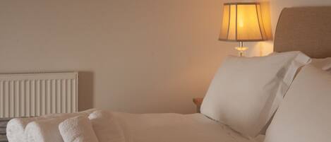 A neatly made bed with white linens and two rolled towels, a bedside lamp, and a radiator on the wall.