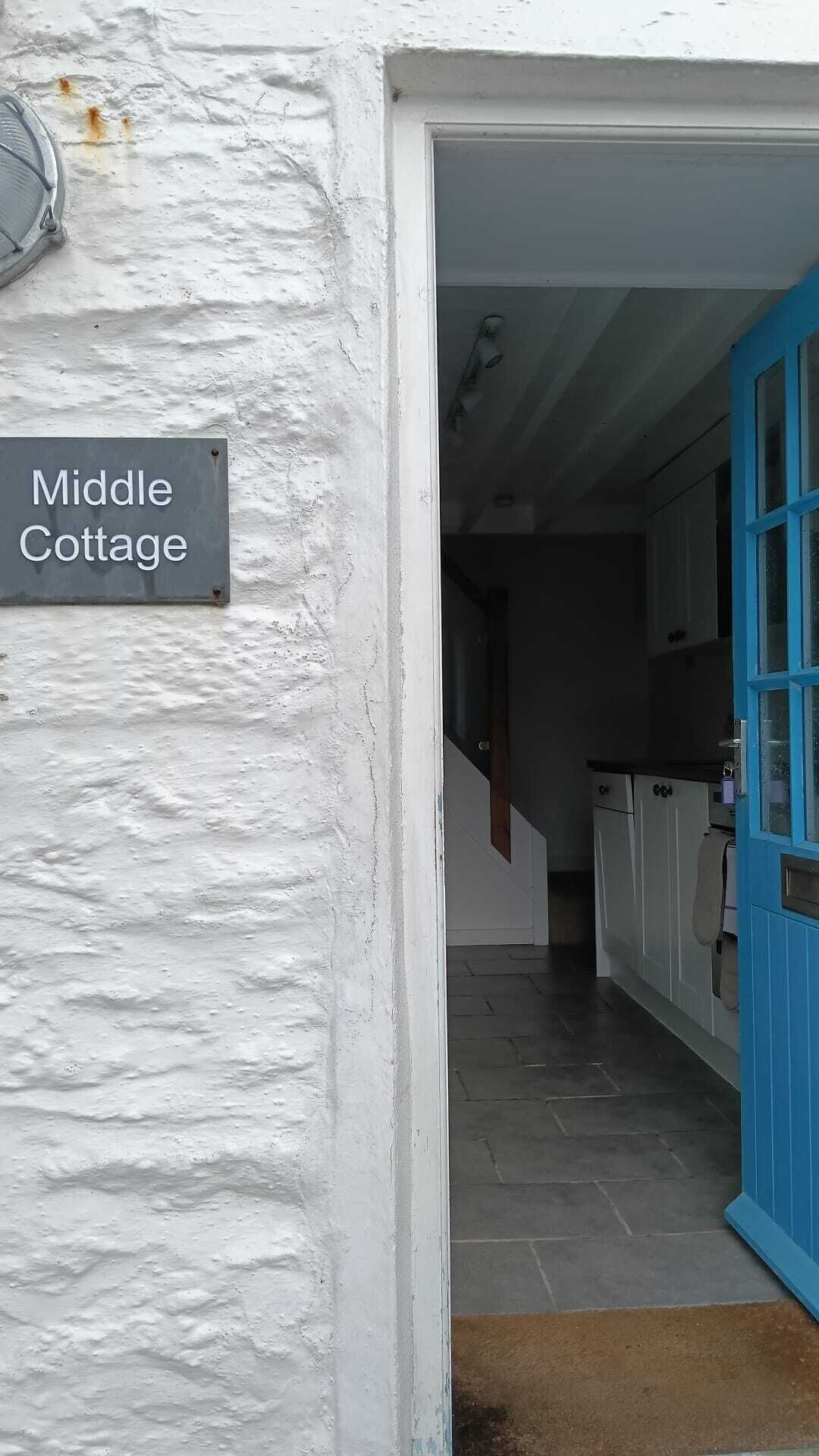 Un mur extérieur blanc avec une pancarte indiquant « Middle Cottage », une porte bleue et un espace cuisine comprenant des placards et un plan de travail.