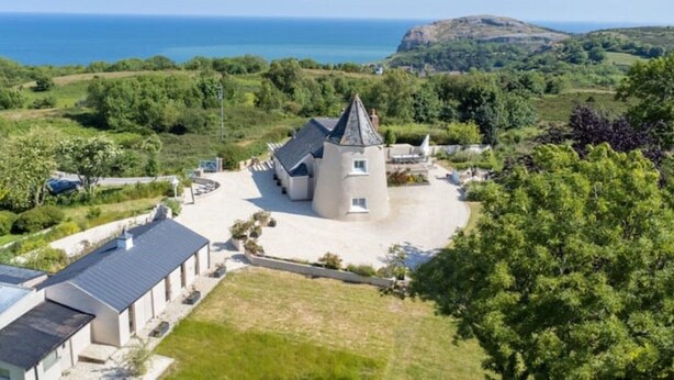 Ein Gebäude mit einem zentralen Turm, umgeben von einem befestigten Bereich und Grünflächen, mit Blick auf eine Küstenlandschaft.