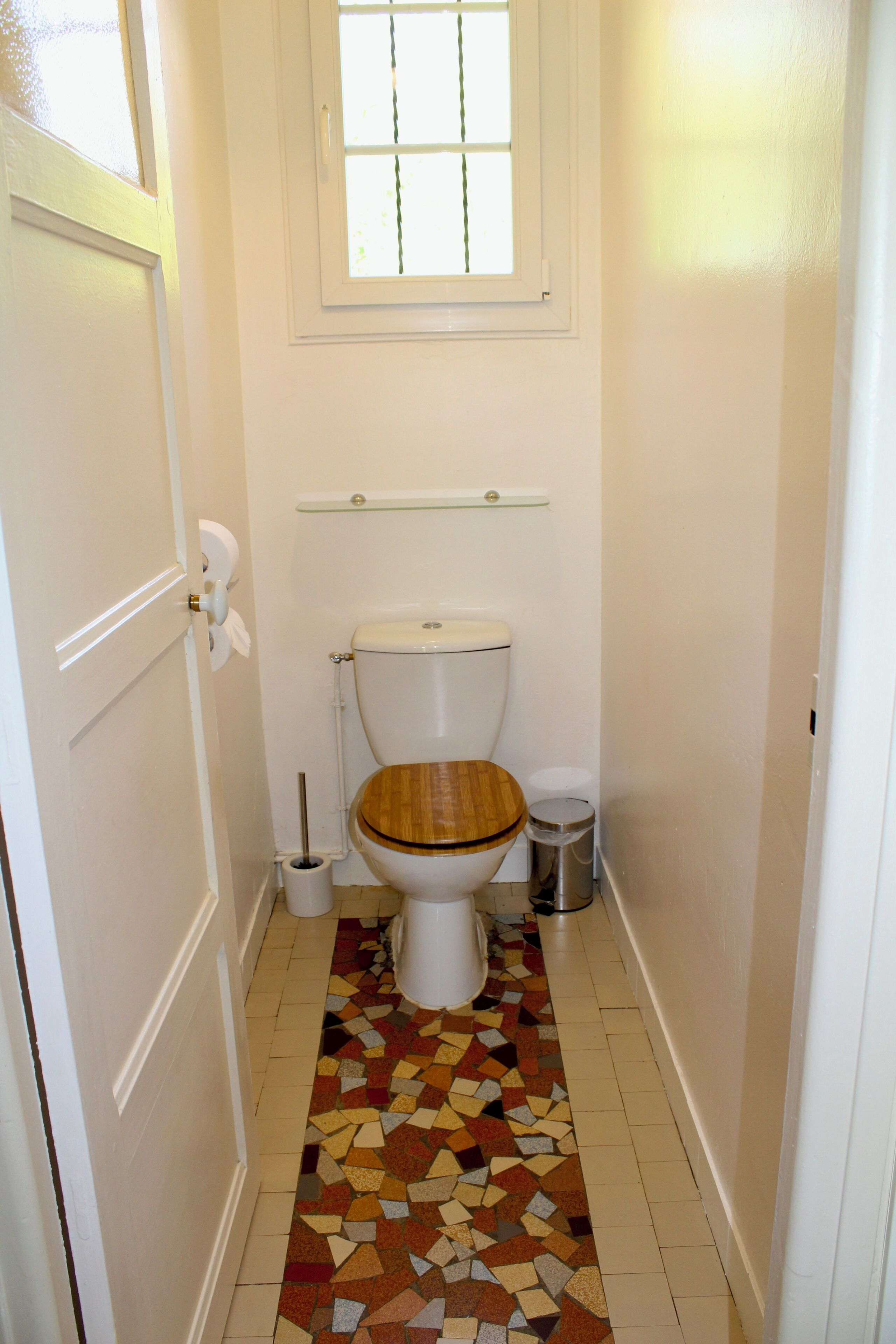 A small bathroom with a wooden toilet seat, a white toilet, a glass shelf above the toilet, a tiled floor with a colorful pattern, and a white door with a doorknob.