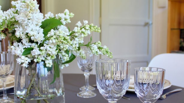 Een gedekte eettafel met helder glaswerk en een vaas witte bloemen.